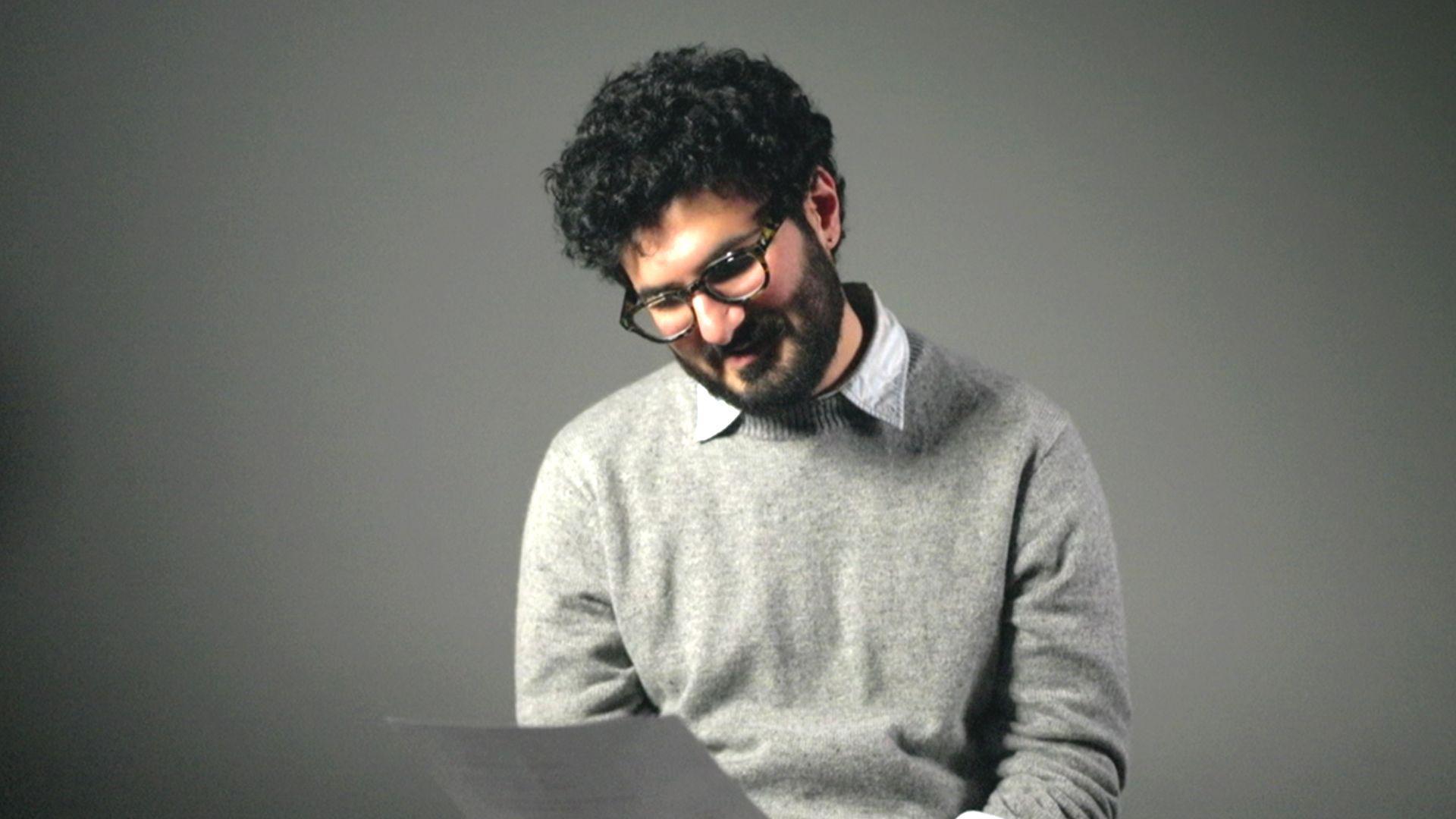 Armen Aramjan sedi u studiju, na stolici i gleda u dokumenta koja drži u desnoj ruci, ima tamnu kobvrdžavu kosu, naočale, svetlo sivi džemper i belu košulju ispod