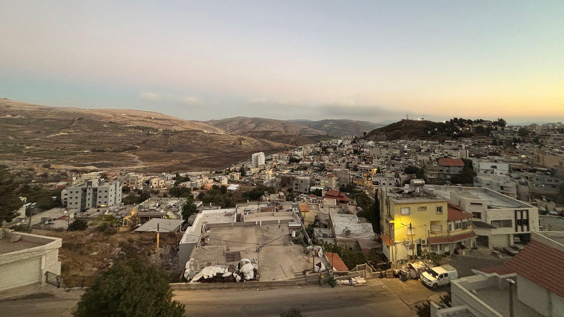 Pogled na mali grad, sa uglavnom niskim betonskim zgradama. U pozadini su brda. Oblačno nebo je plavkasto-sivo sa bledim žutim nagoveštajem sunčevih zraka na horizontu.