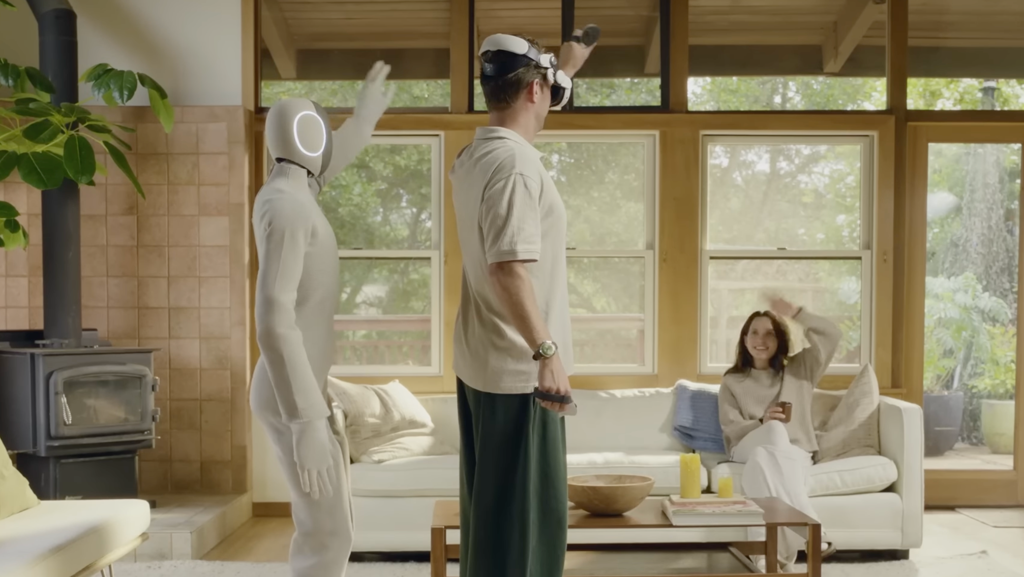 Robot pored muškarca koji je podigao ruku, dok žena sedi na krevetu