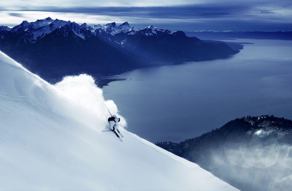 Skijaš se probija kroz svež sneg na strmoj planinskoj padini, stvarajući oblak snežnog praha.