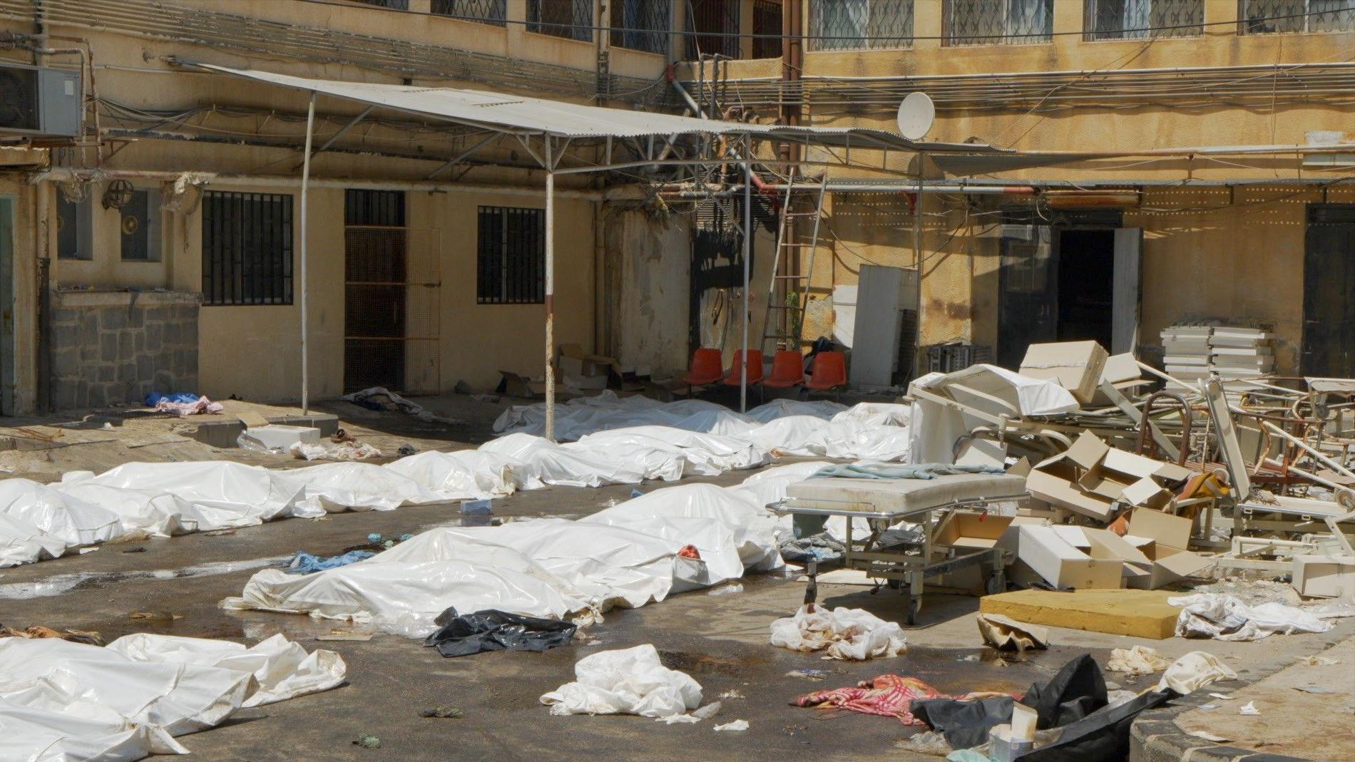 Bele plastične folije prekrivaju tela na parkingu bolnice u Suveidi. Ostaci polomljenih stolica i bolničkih kreveta takođe su razbacani po zemlji.