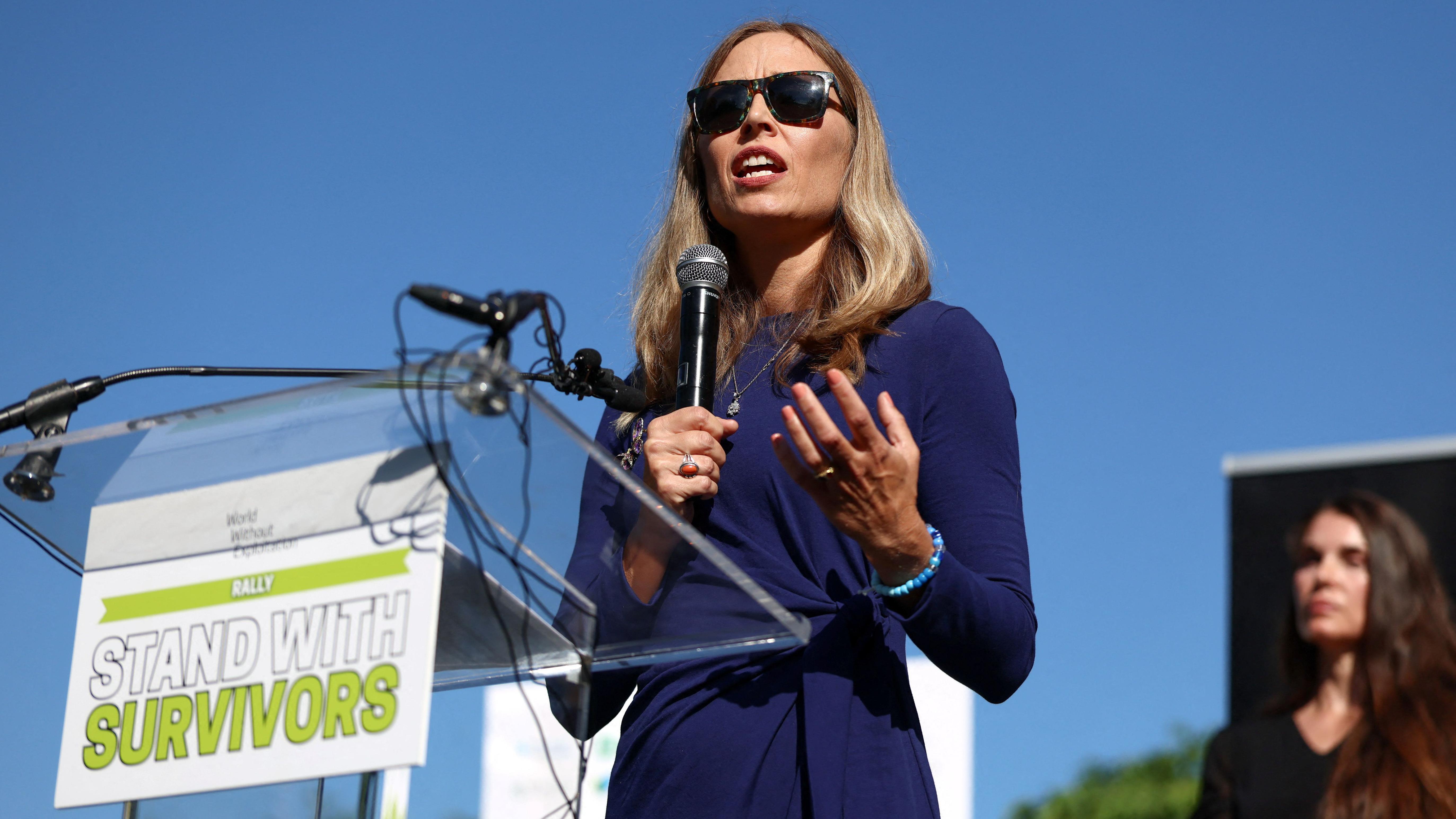 Eni Farmer, jedna od Epstinovih žrtava, stoji za podijumom sa mikrofonom u ruci, nosi naočare za sunce i svečanu plavu haljinu. Sunčano je i na podijumu je moto „Stanite uz preživele“ napisan na tabli okrenutoj ka publici.