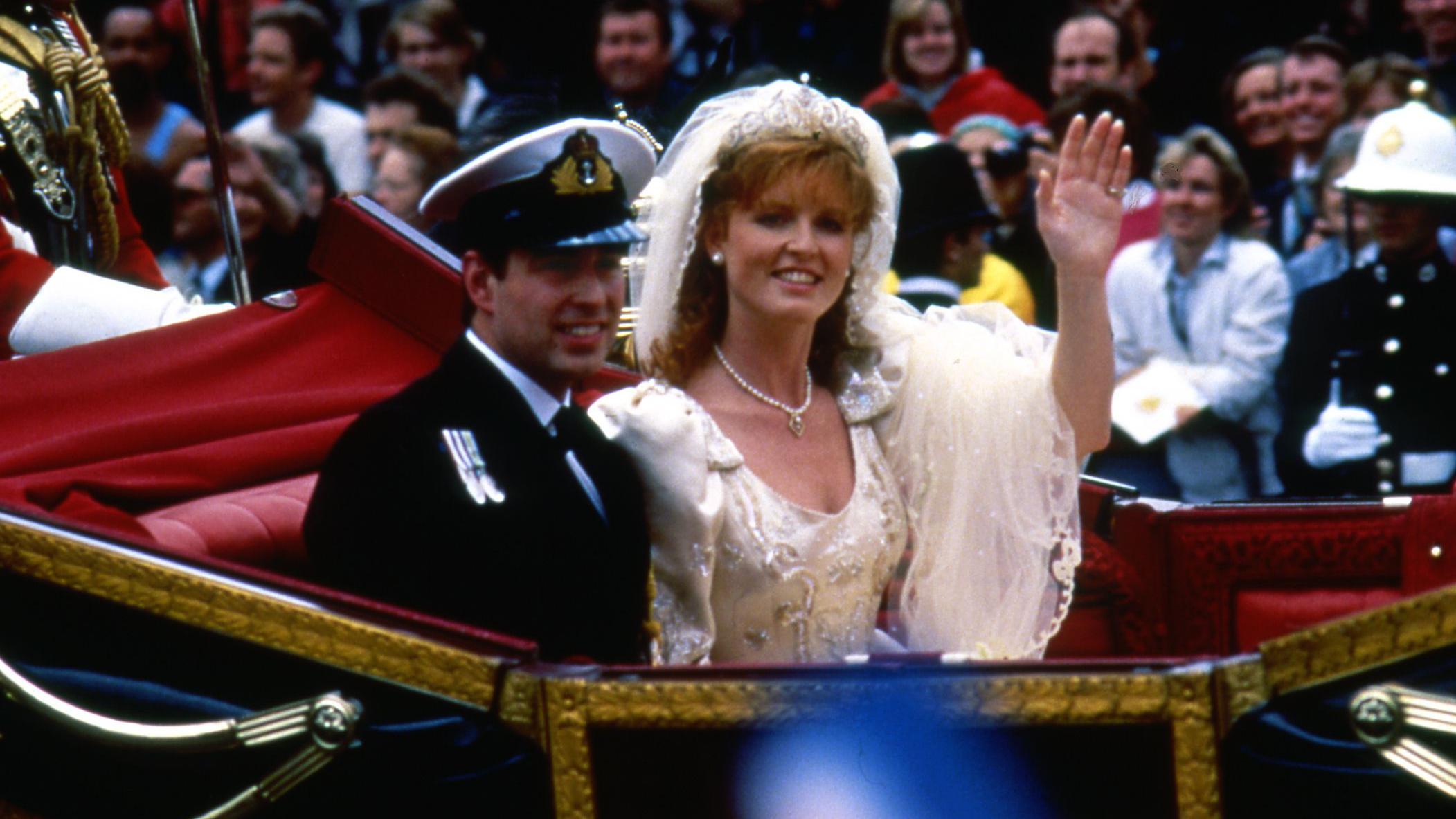 Andrew in a naval uniform alongside Sarah Ferguson in a white wedding dress riding in an open-topped carriage. Ms Ferguson is waving. Crowds line the street behind them.