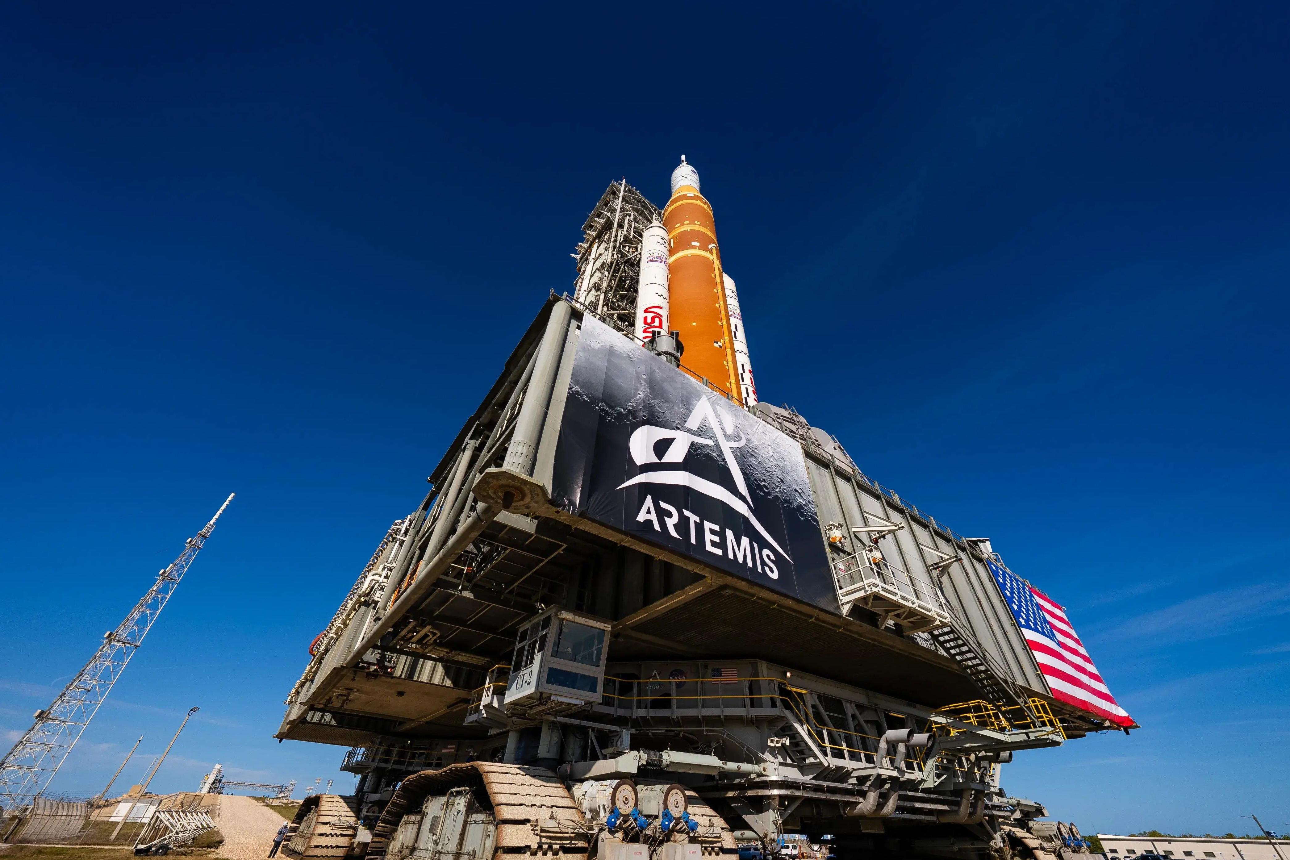 Nasin ogromni sistem za lansiranje u svemir i letelica Orion u svemirskom centru Kenedi na Floridi naspram vedrog plavog neba sa zastavama Artemide i američkom zastavom 