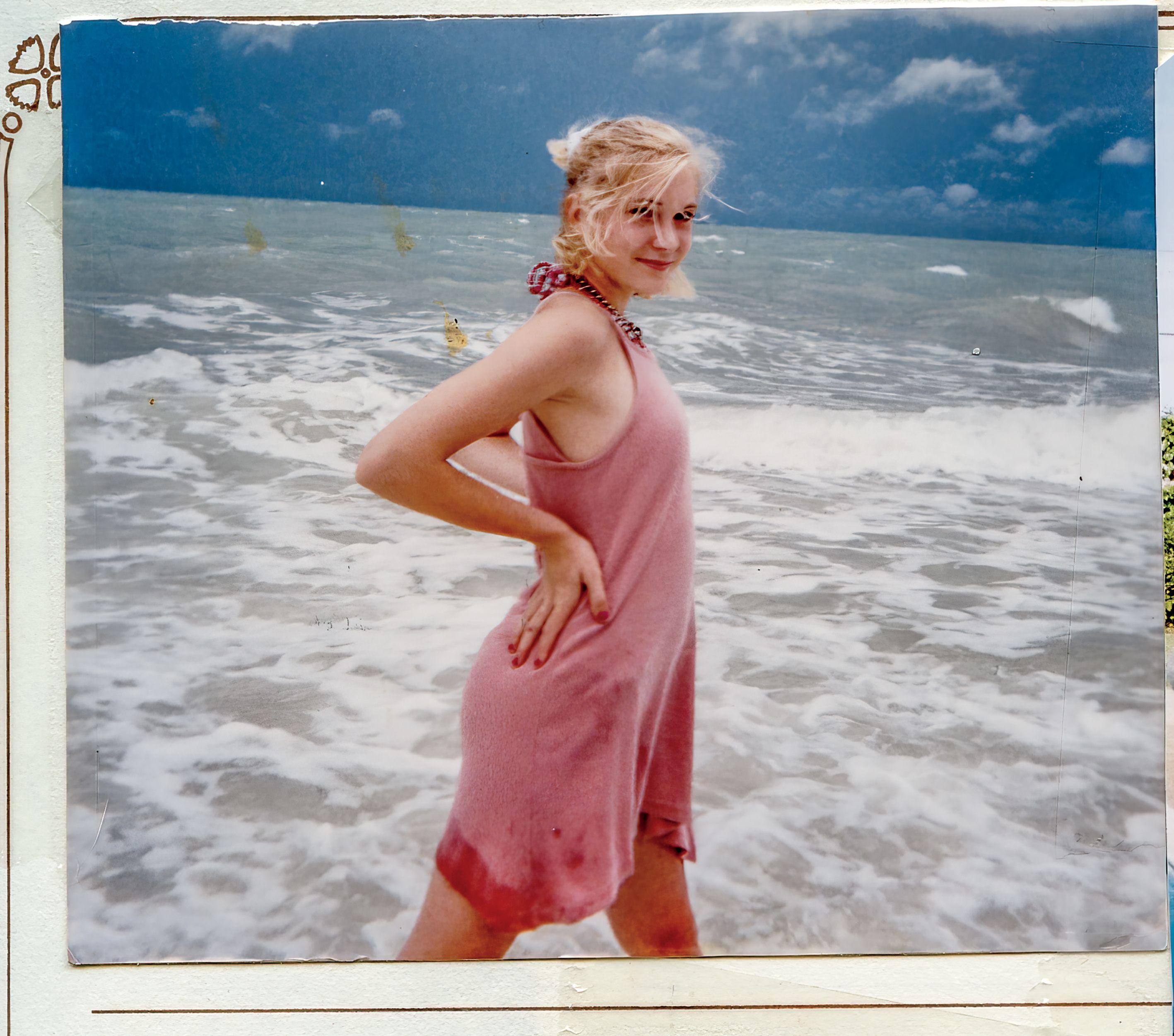 VIctoria Guiffre standing in the sea, wearing a pink dress with splashes on the lower part. Her hands are held to her hips. She is looking at the camera with hair tied back. The sea is grey and there are clouds in the sky. The photo is within a photo binder.