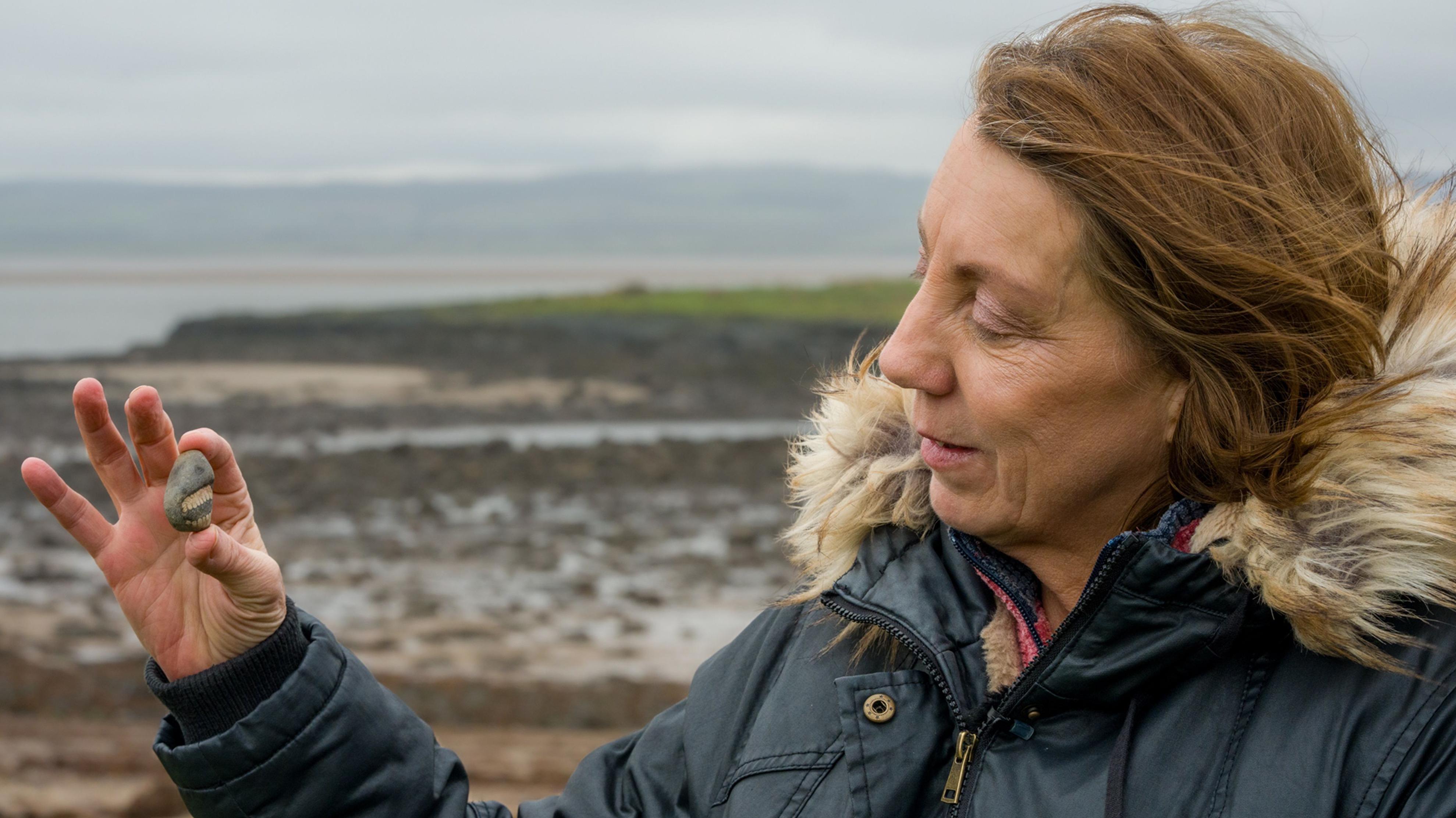 Žena sa smeđom kosom do ramena u plavom kaputu sa krznenom kapuljačom, u desnoj ruci drži kamen sa umetnutim fosilom