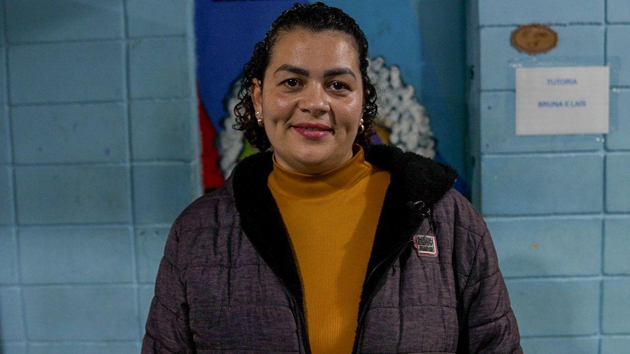 Ana Gabriela Lima u zimskoj jakni i narandžastoj rolci, nasmejano gleda u objektiv kamere