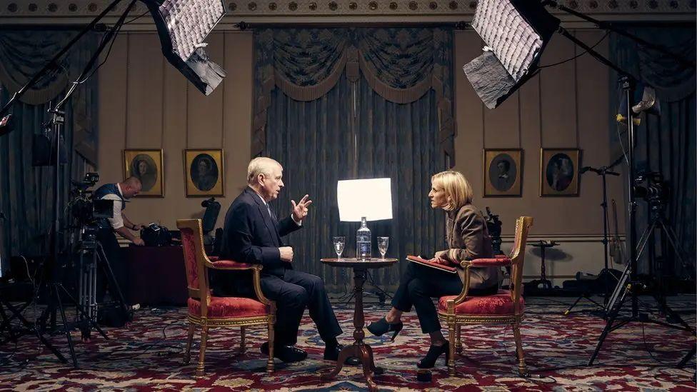 A side shot of Andrew speaking to Emily Maitlis during the recording of the 2019 BBC Newsnight interview reported worldwide. Large lights on large stands tower above them and another light is at their head level, in the centre of a window bay with large blue curtains drawn. A man wearing a waistcoat is in the corner of the shot, tending some equipment. A table with two glasses and a bottle of water is between Maitlis and Andrew.