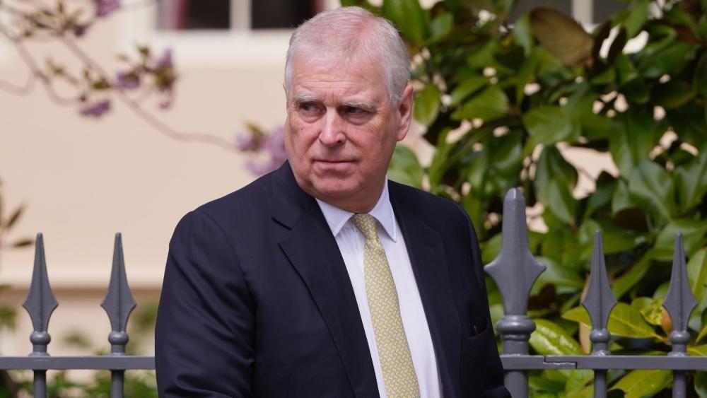 Andrew walking looking solemn in front of a metal grey fence which has some foliage behind it. You can only see from his chest up. He wears a black blazer, a gold tie and white shirt 