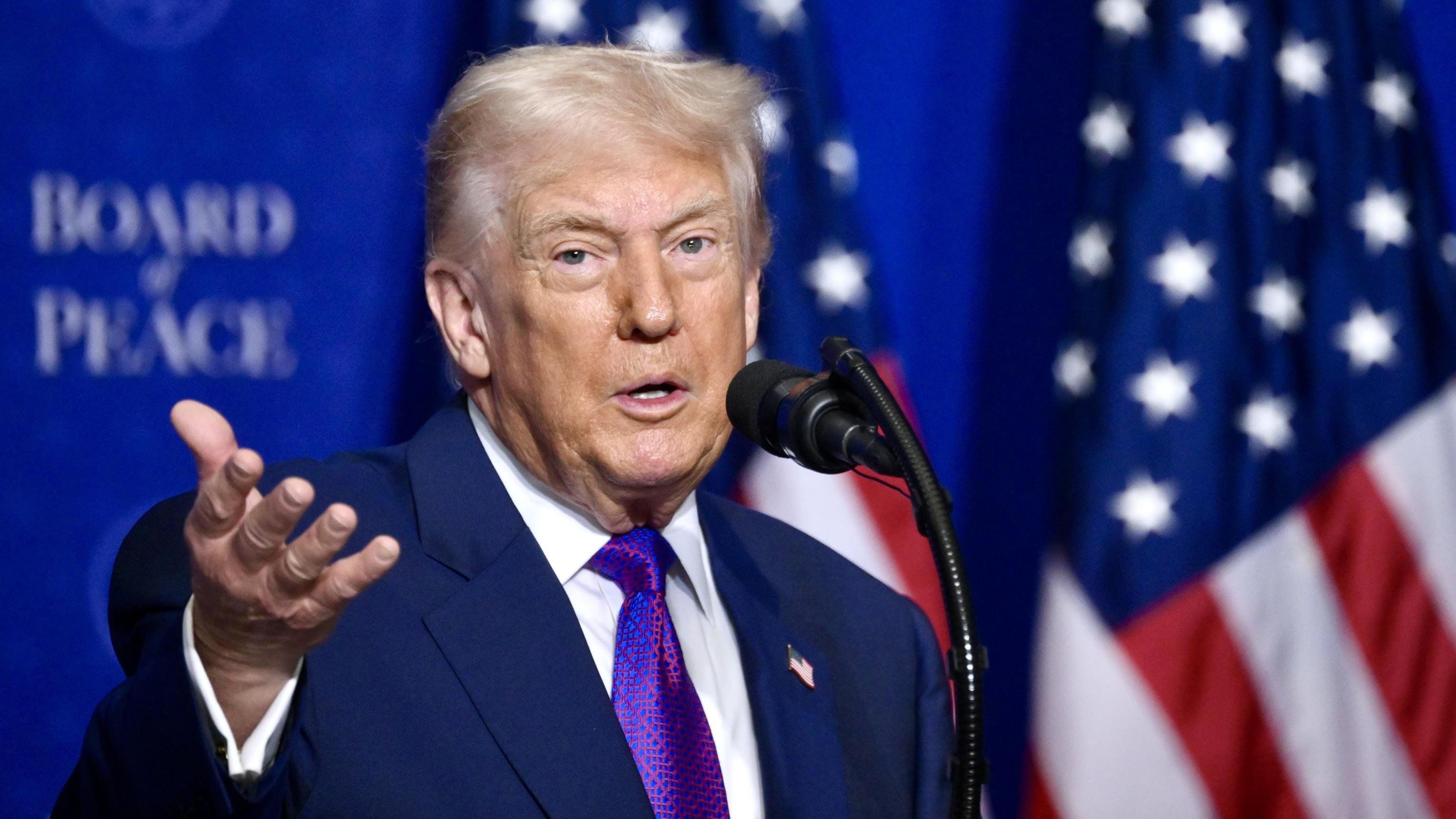 Tramp govori tokom sastanka Odbora za mir u Vašingtonu 19. februara 2026. Drži desnu ruku podignutu dok govori u mikrofon, sa američkom zastavom iza sebe.