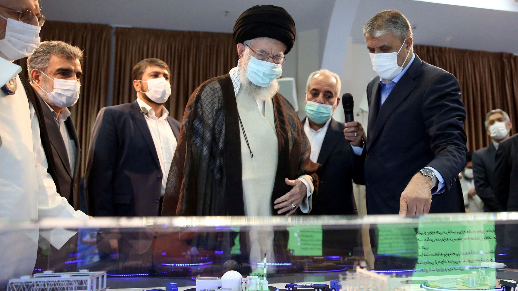 Ajatolah Hamnei: Kako je iranski vrhovni vođa držao vlast čeličnim stiskom 18 Khamenei views a model of a nuclear facility, in Tehran on 11 June 2023. He is wearing a blue face mask and traditional clothes while other men stand around him. One is wearing a white laboratory jacket. The model is in a display cabinet and consists of a white reactor dome and other buildings in one location, along with the framework of a rectangular building with models of people in it nearby.
