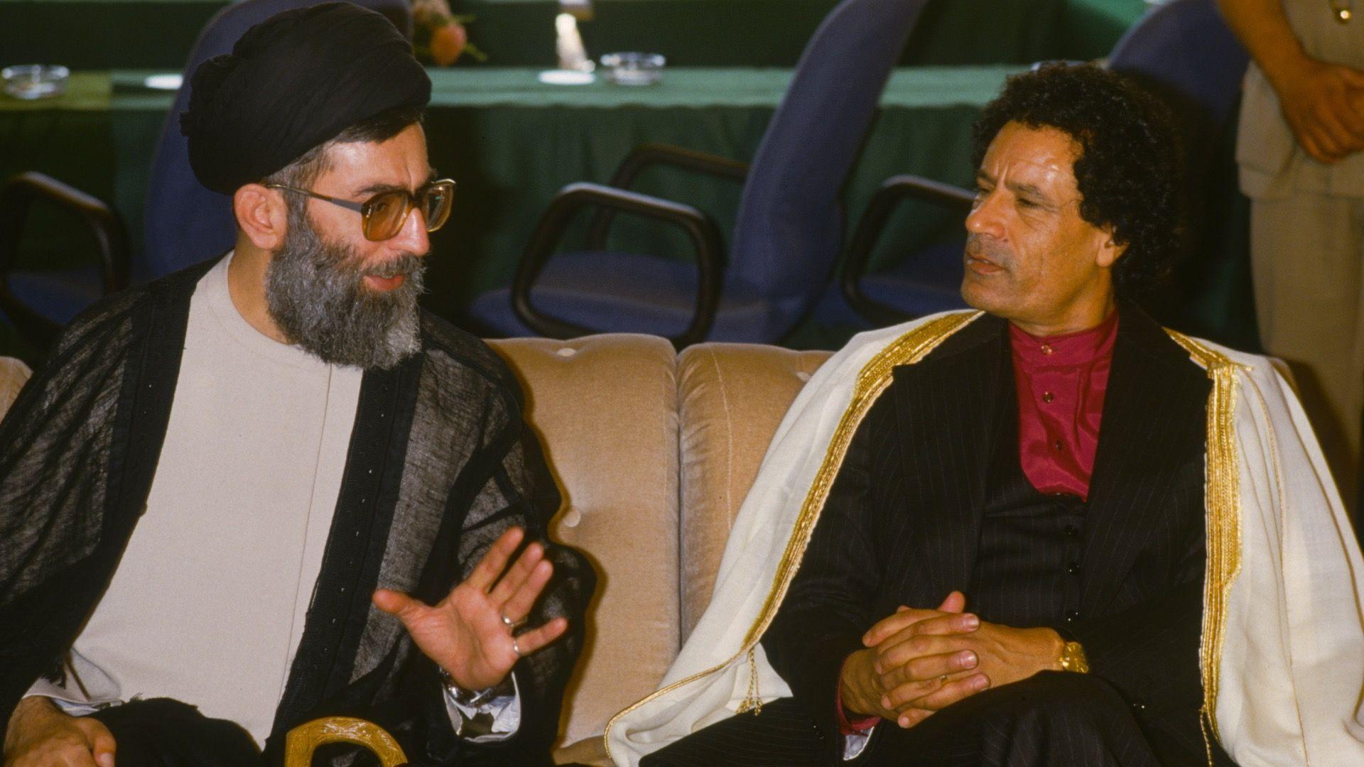 Ajatolah Hamnei: Kako je iranski vrhovni vođa držao vlast čeličnim stiskom 10 Gaddafi , sitting to the right of the image, looks at Khamenei as the Iranian leader talks. Gaddafi is holding his hands together on his lap. He is wearing a white robe with golden embroidery on the sides, a black jacket and dark red shirt with no collar, buttoned to the top. Khamenei is holding his hand up as he speaks. He is wearing black clothing on his head, a grey and black jacket and cream shirt. The pair are sitting next to each other on a sofa, with desk chairs and a meeting table behind them.