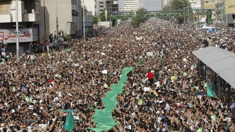 Ajatolah Hamnei: Kako je iranski vrhovni vođa držao vlast čeličnim stiskom 12 A wide city street packed with thousands of male and female protests on 15 June 2009. Some people are holding green flags, while others are holding posters. Tall buildings are on either side and a metal bridge is in the background.