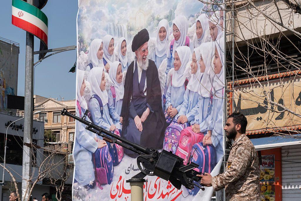 Iran ne može vojno da pobedi Ameriku, ali evo kako njegove vođe planiraju da opstanu 4 Mušarac u vojničkoj uniformi drži oružje u ruci iza njega plakat ubijenih devojčica i iranskog vođe