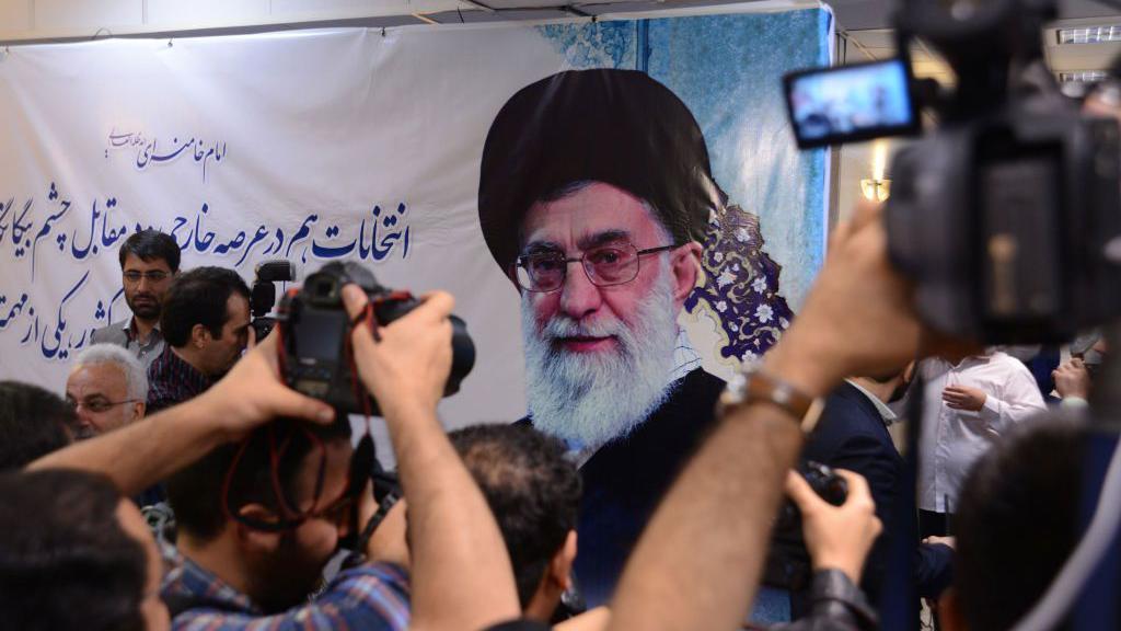 Ajatolah Hamnei: Kako je iranski vrhovni vođa držao vlast čeličnim stiskom 11 Ayatollah Khamenei shown on a white-cloth banner with Persian writing drawn on it. Cameras with video screens are pointed towards the banner.