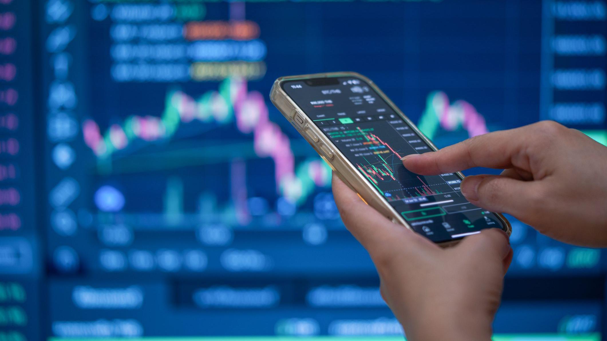 Jesu li Trampove neizgovorene reči uticale na cenu nafte 1 A trader's hands using their phone to make a trade in front of a trading screen in soft focus