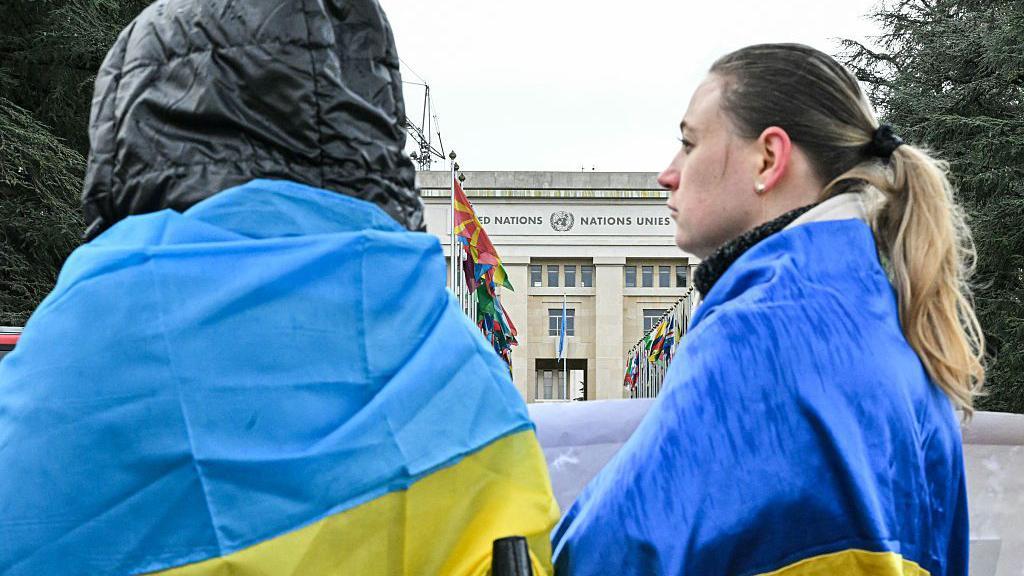 Mirovni pregovori u zastoju, Rusija tvrdi da je zauzela ceo Lugansk 3 Dvoje ljudi leđima okrenuti kameri ispred zgrade Ujedinjenih nacija u Ženevi ogrnuti ukrajinskim zastavama