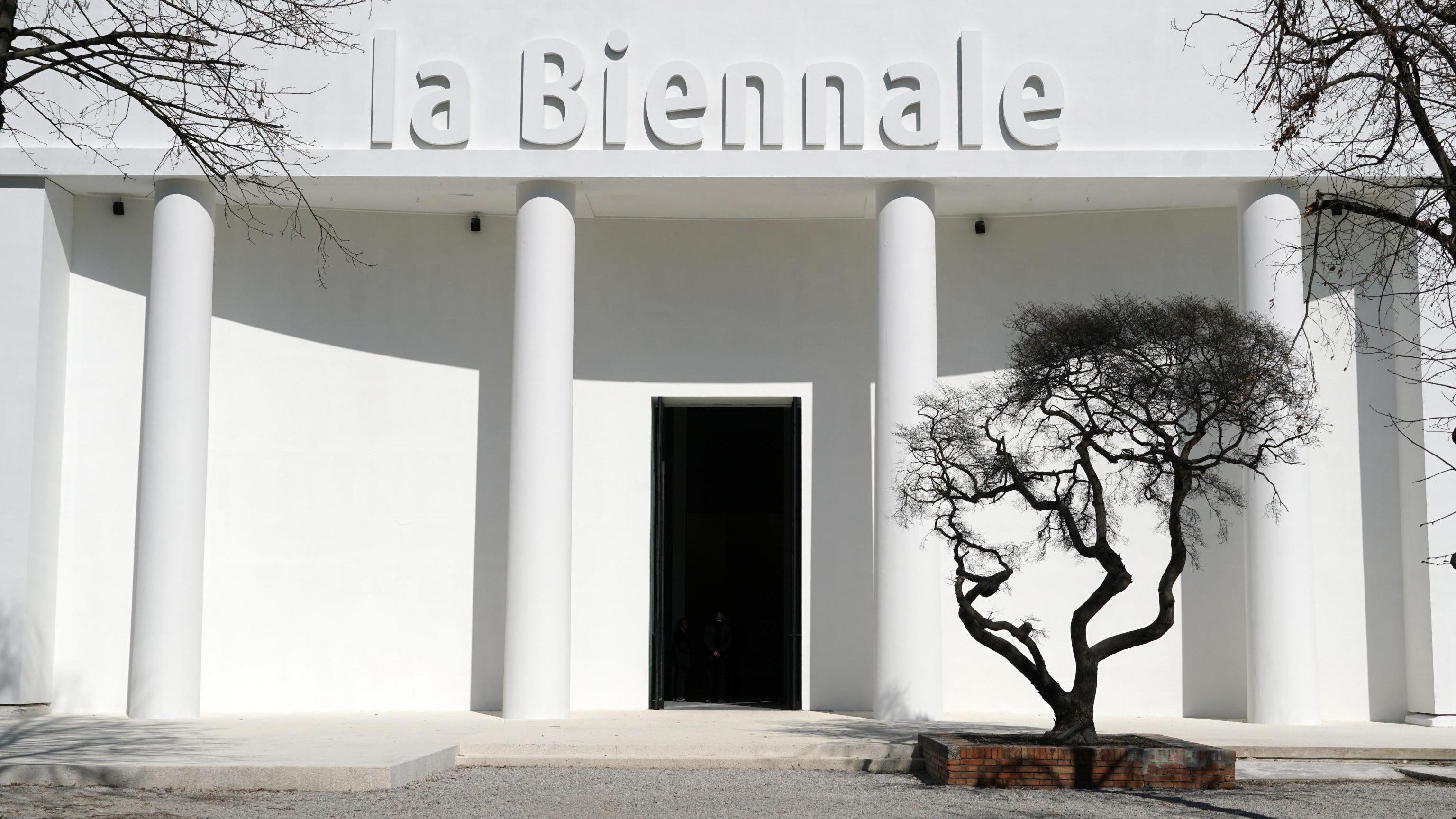 Bela zgrada sa natpisom „la Biennale“ i crnim drvetom ispred nje