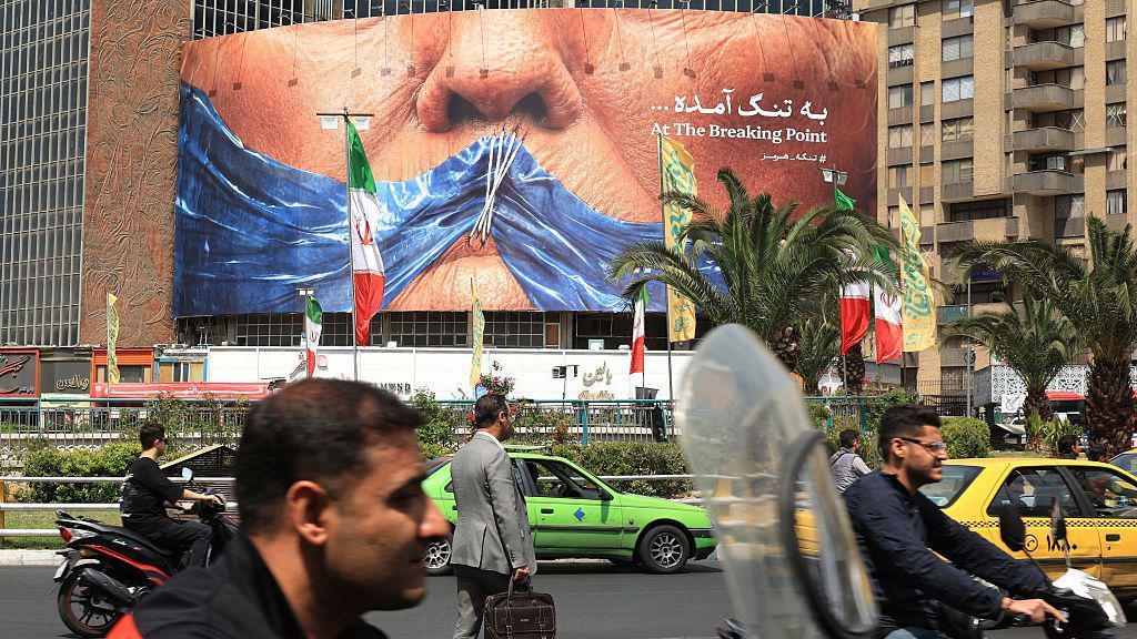 Američko-izraelski sukob sa Iranom: Jedan rat, dve tvrdnje o pobedi 1 Stanovnici Irana prolaze automobilima, taksijima, motorima, i pešice pored antiameričkog bilborda koji se odnosi na američkog predsednika Donalda Trampa i Ormuski moreuz. U pozadini se vide iranske zastave, palme i zgrade