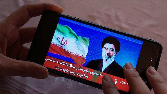 Tajna mreža krijumčari Starlinkovu tehnologiju u Iran kako bi pobedila blokadu interneta 2 ruke drže mobile telecom na kome je fotografija vođe Modžatbe Hamneija