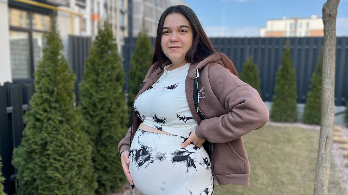 Trudna žena stoji u bašti sa rukom na stomaku. Ima dugu, ravnu, tamnu kosu i nosi odgovarajući top i pantalone koje su bele sa nekoliko crnih mrlja. Takođe ima svetlo smeđu duksericu.