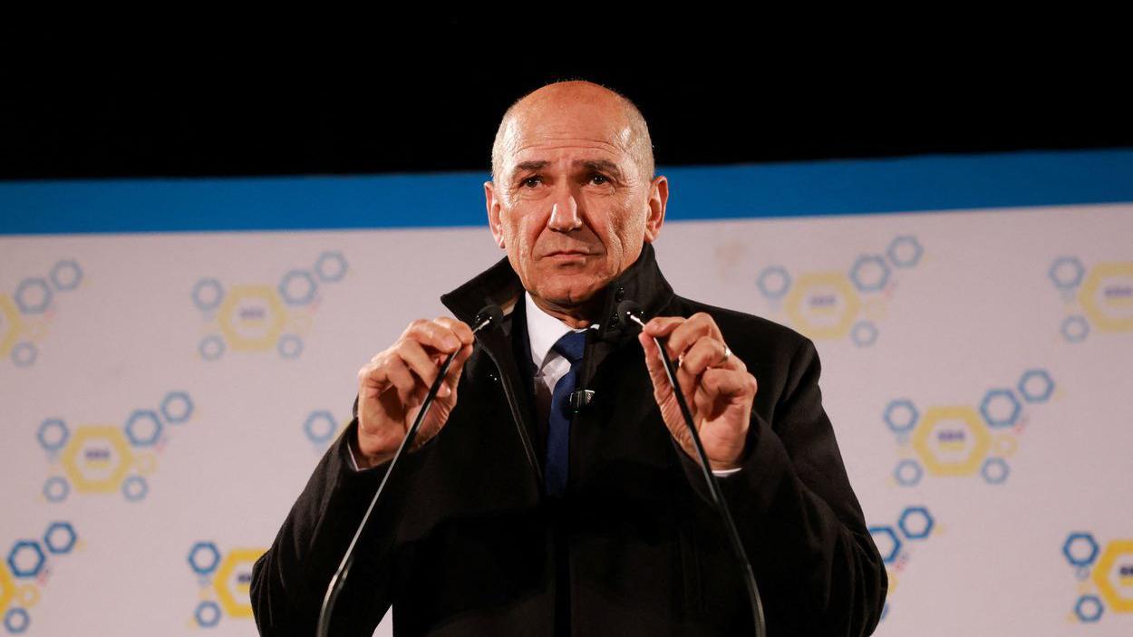 Janez Janša: Od komunističkog disidenta do konzervativnog premijera Slovenije 2 Janez Janša u crnom kaputu za govornicom, rukama drži dva mikrofona