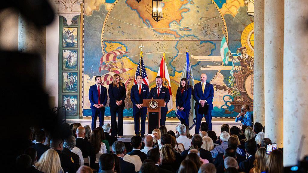 Amerika optužila kubanskog lidera Raula Kastra za ubistvo zbog obaranja dva aviona 1996. 2 Džejms Utmajer, Madelin Pumarijega, Tod Blanš, Džejson Reding Kinjones, senator Ešli Mudi i Kristofer Raja, svi u odelima, stoje rame uz rame ispred podijuma na bini. Gomila se okupila ispred njih.