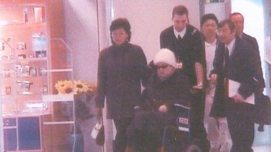 Zašto Kim Džong Un nikad ne govori o majci 7 Ko Jong Hui, sa naočarima za sunce i belom kapom, sedi u invalidskim kolicima koja gura muškarac. Nekoliko ljudi je oko nje u bolnici u Francuskoj.