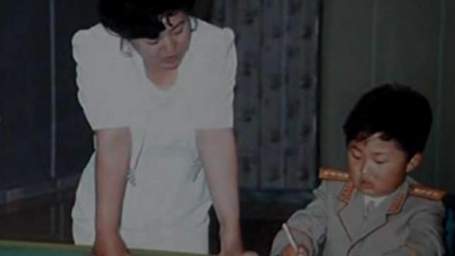 Zašto Kim Džong Un nikad ne govori o majci 5 Ko Jong Hui, u beloj haljini, stoji s leve strane. Mladi Kim Džong Un, u vojnoj uniformi, sedi i piše olovkom.