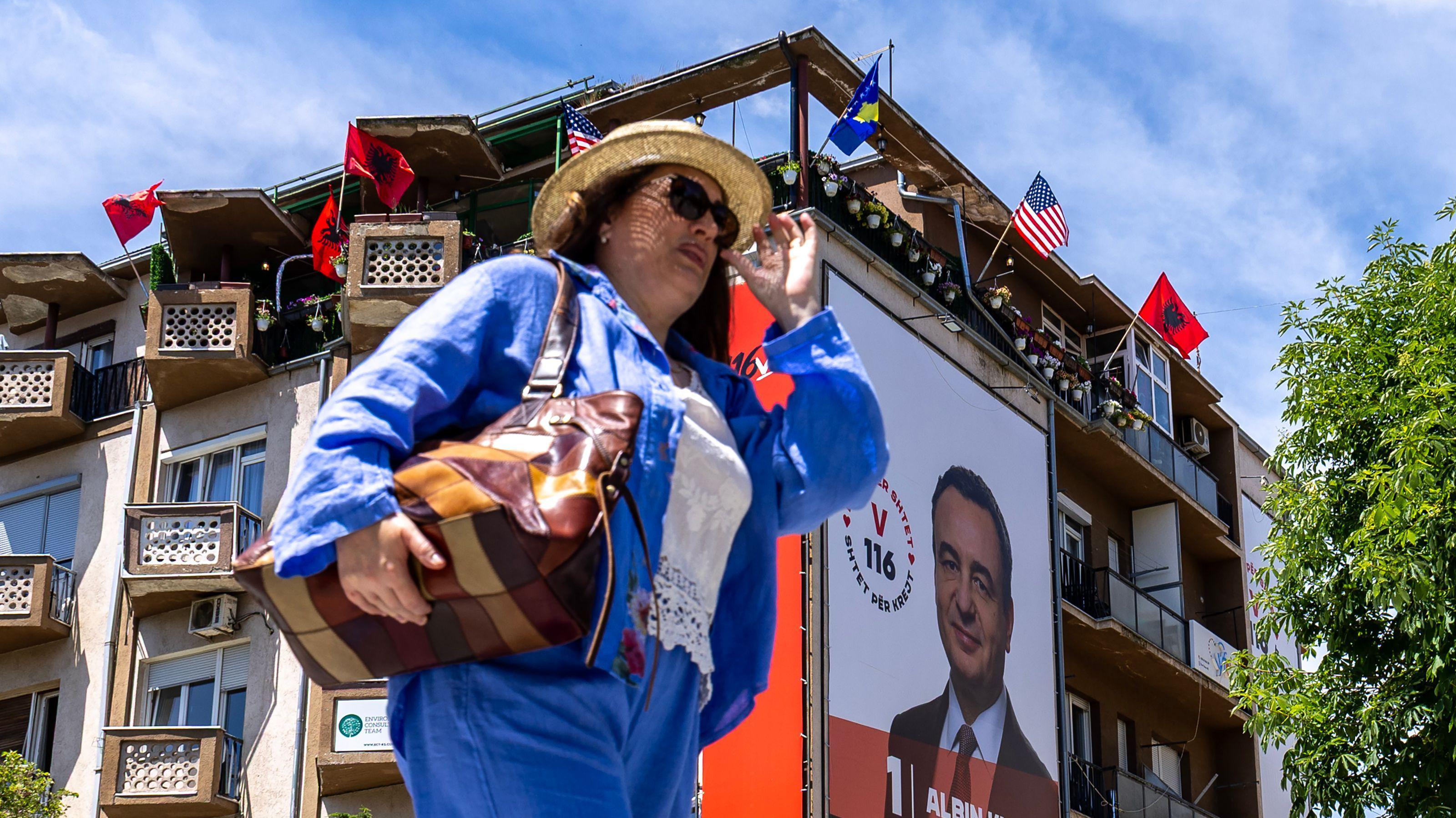 Kosovski politički meč bez kraja: 'Kurti ima brojke, ali ne i političku volju' 1 Žena ide ulicom sa naočalama za sunce iza nje plakat premijera Kosova Aljbina Kurtija i zastave Kosova, Amerike i Albanije