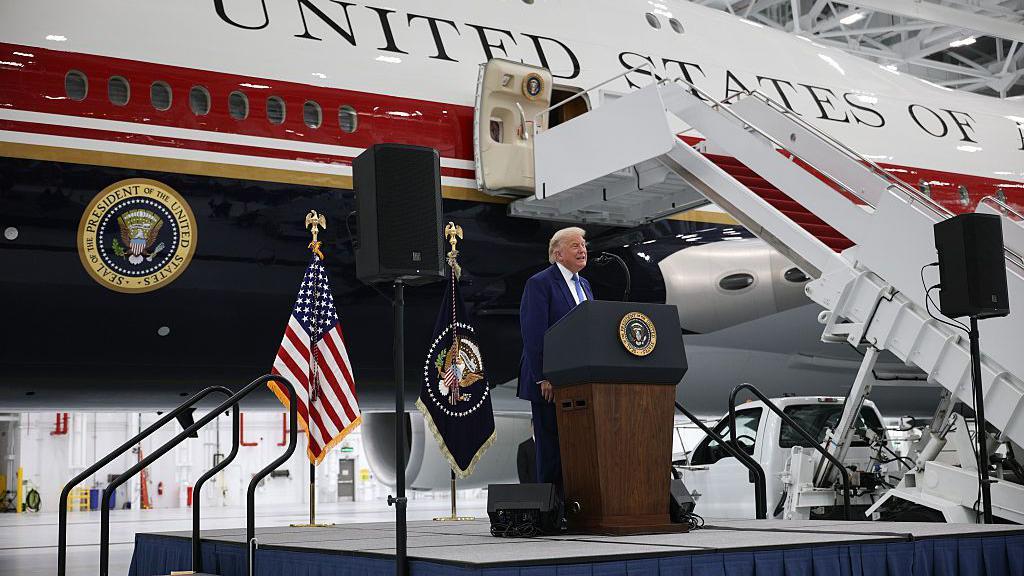 Tramp predstavio luksuzni avion, poklon Katara, koji postaje deo predsedničke flote 1 Trump standing at a podium on a stage in front of the new jet, painted white with dark red and blue stripes. There is a presidential seal and the words "United States of" visible.