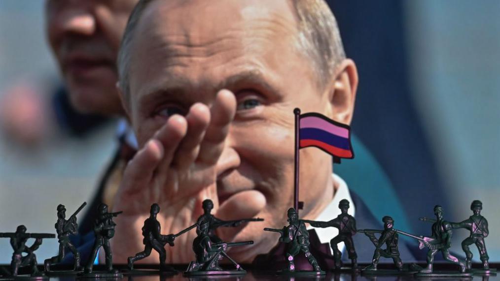 U pozadini je krupni plan fotografije predsednika Putina, koji maše rukom, u prvom planu su mnoštvo tamnozelenih/crnih vojnika igračaka i ruska zastava (sa horizontalnim prugama belim (gore), plavim i crvenim (dole).